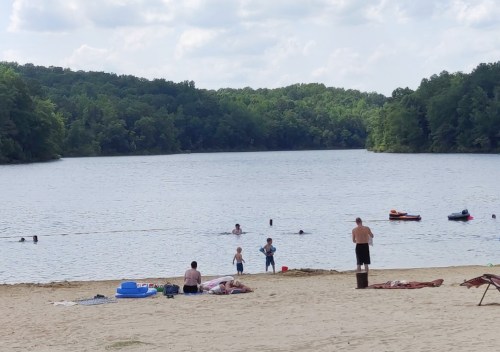 A sandy beach by a lake with people swimming, playing, and relaxing under a partly cloudy sky.