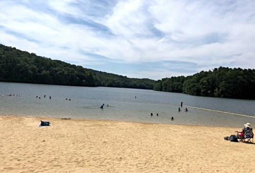 A sandy beach by a calm lake, with people swimming and relaxing under a blue sky with scattered clouds.