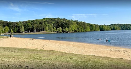 A serene lakeside view with a sandy beach, green trees, and people enjoying the water on a sunny day.