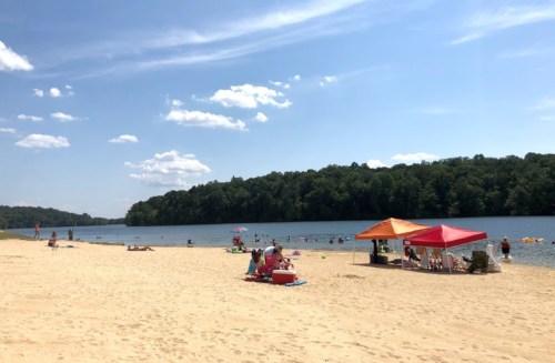 A sandy beach by a calm lake, with people relaxing, swimming, and colorful tents under a clear blue sky.