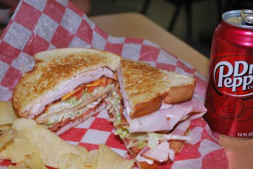 A toasted sandwich with turkey, lettuce, and tomato, served with chips and a can of Dr Pepper on a checkered paper.