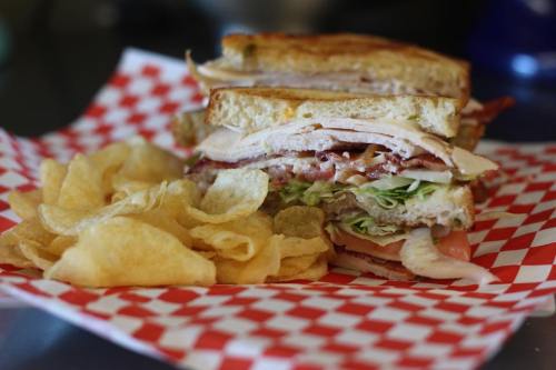 A stacked sandwich with turkey, lettuce, and bacon, served with a side of potato chips on a red checkered paper.