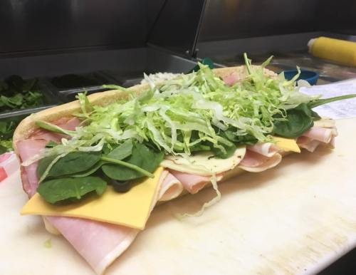 A large sub sandwich filled with layers of ham, cheese, spinach, and shredded lettuce on a cutting board.