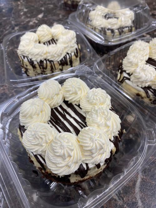 Several individual cakes topped with whipped cream and chocolate drizzle, displayed in clear plastic containers.