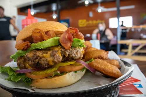 A close-up of a juicy burger topped with bacon, lettuce, tomato, pickles, and cheese, served with crispy potato wedges.