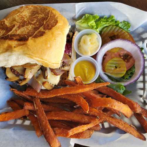 A sandwich with grilled onions and potatoes, served with sweet potato fries, lettuce, tomato, pickles, and condiments.