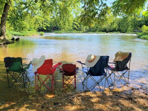 Five colorful camping chairs with wide-brimmed hats sit by a calm river, surrounded by lush greenery.
