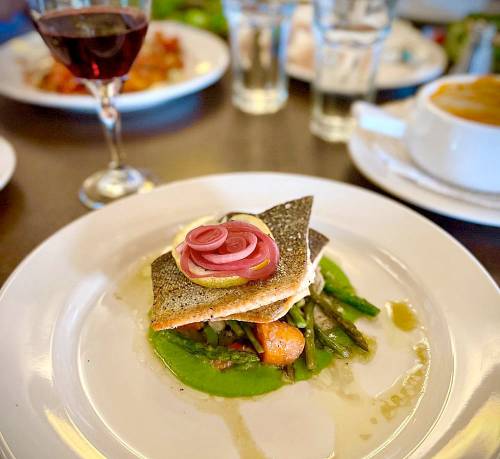 A beautifully plated fish dish with pickled onions, served on a bed of green vegetables, alongside a glass of red wine.