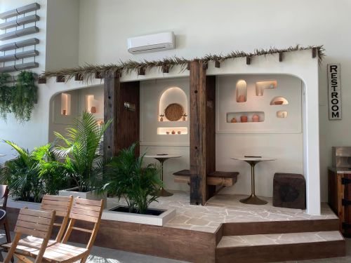 A modern café interior featuring plants, wooden accents, and decorative shelves with soft lighting. Restroom sign visible.