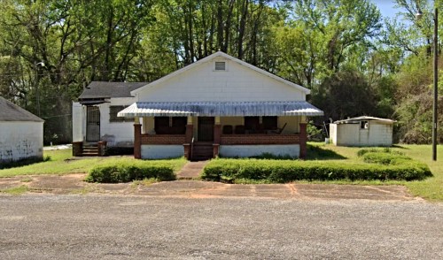 A white house with a porch, surrounded by greenery, and a small outbuilding in the background.