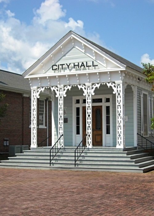 Historic city hall building with a white facade, decorative porch, and steps leading to the entrance. Blue sky in the background.