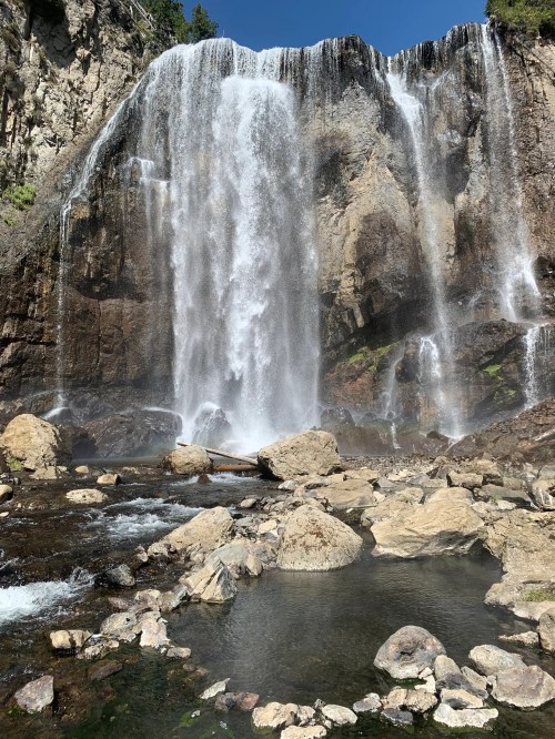 A majestic waterfall cascading over rocky cliffs into a serene pool, surrounded by lush greenery and clear blue skies.