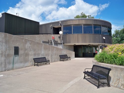 Modern building with large windows, benches in front, and a landscaped area under a blue sky with clouds.