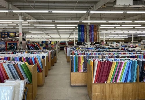 A spacious fabric store filled with colorful rolls of fabric arranged on wooden shelves. Brightly lit with high ceilings.