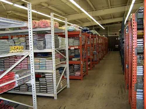A spacious warehouse aisle lined with shelves stacked with neatly organized blankets and textiles.