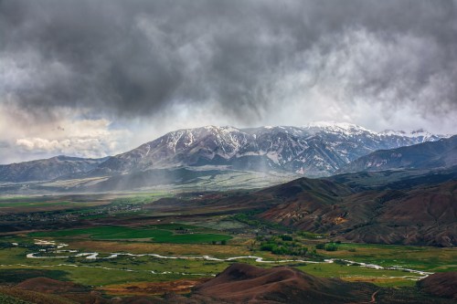 A dramatic landscape featuring snow-capped mountains, lush green valleys, and dark, cloudy skies.