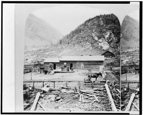 A historic black-and-white photo of a log cabin in a mountainous landscape, with people and a horse in the foreground.