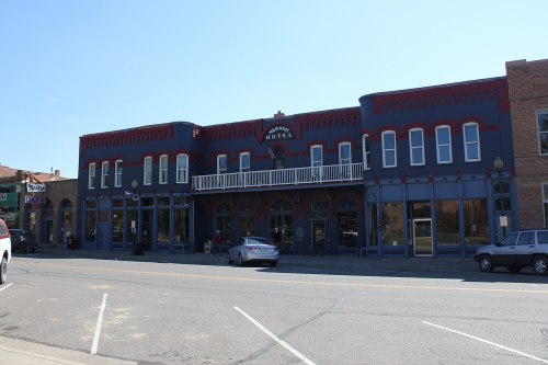 Historic hotel building with red and blue accents, featuring a balcony and large windows, set on a street with parked cars.