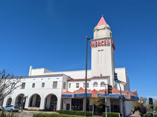 Historic Merced Theatre with a tall tower and red signage against a clear blue sky. Green traffic lights in view.