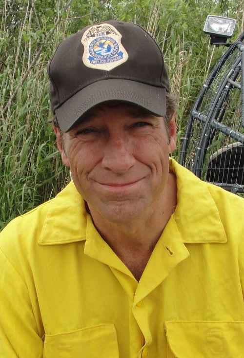 A man in a yellow shirt and cap smiles, sitting in a grassy area near an airboat.