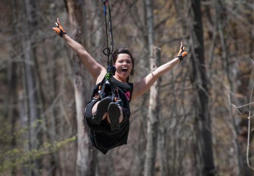 A person joyfully zip-lining through a forest, arms raised and smiling, surrounded by trees.