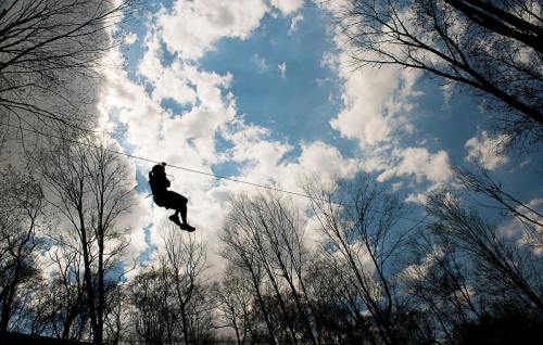 A person zip-lining through a forest against a backdrop of clouds and blue sky.