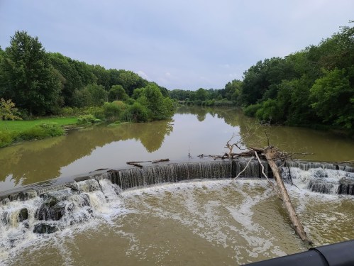 A serene river scene featuring a small waterfall, surrounded by lush greenery and a cloudy sky.