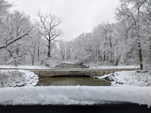 A snowy landscape featuring a river and a bridge, surrounded by trees blanketed in white snow.