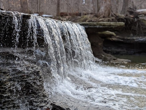 A close-up of a small waterfall cascading over rocky ledges into a serene pool, surrounded by trees.