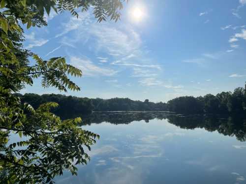 A serene lake reflects the sky and trees, with the sun shining brightly above and wispy clouds in the blue sky.