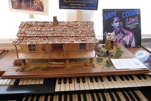 A detailed model of a wooden house with a shingled roof, displayed on a piano, alongside a Sam Cooke album cover.