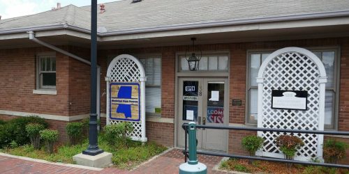 A brick building with a sign reading "OPEN" and decorative latticework, surrounded by greenery and a sidewalk.