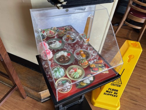 Display case with colorful plastic food replicas, showcasing various dishes, next to a caution sign about slippery floors.