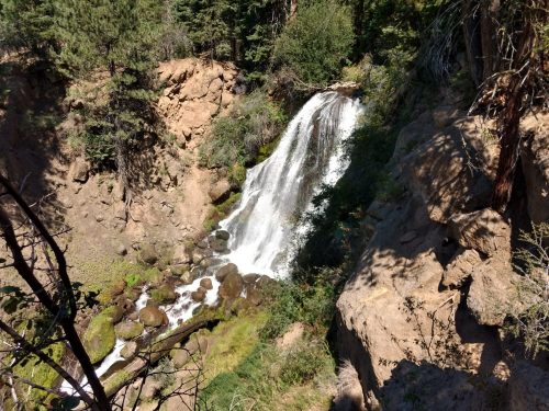 A waterfall cascades down rocky terrain, surrounded by lush greenery and trees in a natural setting.