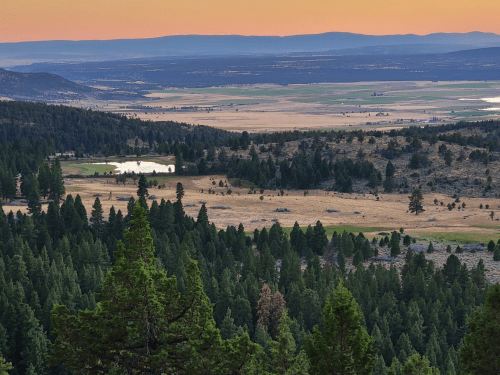 A panoramic view of a valley with trees, fields, and a small pond under a colorful sunset sky.