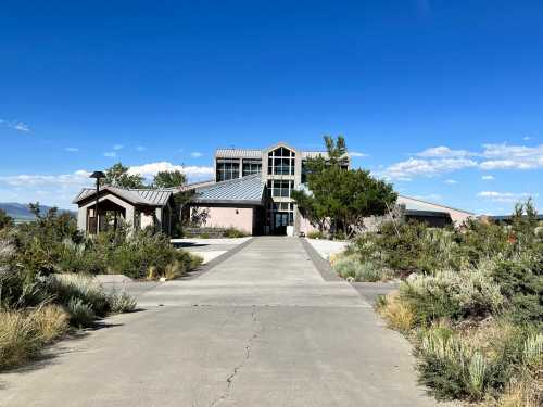 A modern house with large windows, surrounded by greenery and a clear blue sky, with a driveway leading up to it.