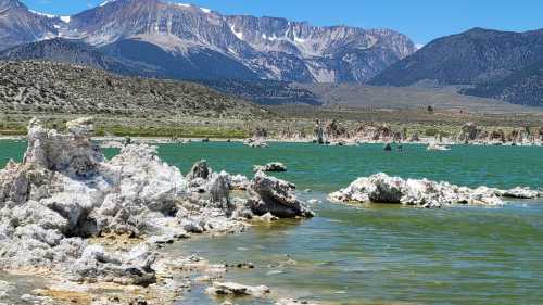 A scenic view of a turquoise lake surrounded by rocky formations and snow-capped mountains under a clear blue sky.