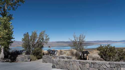 A scenic view of a lake surrounded by mountains, with a person standing near a stone wall and informational signs.