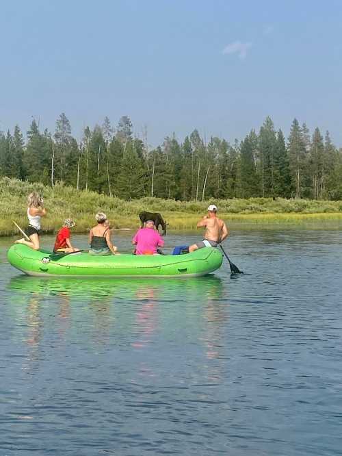 A group of people in a green inflatable boat on a calm lake, surrounded by trees and greenery.