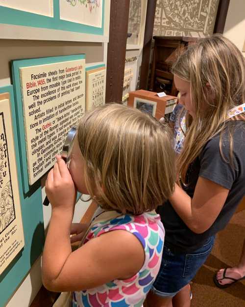 Two children examine a display at a museum, one using a magnifying glass to read a text about historical prints.