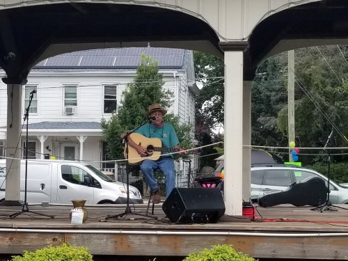 A musician plays guitar and sings on stage under a pavilion, with colorful decorations and a van in the background.