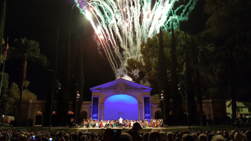 Fireworks illuminate the night sky above an outdoor concert stage filled with musicians and an audience.