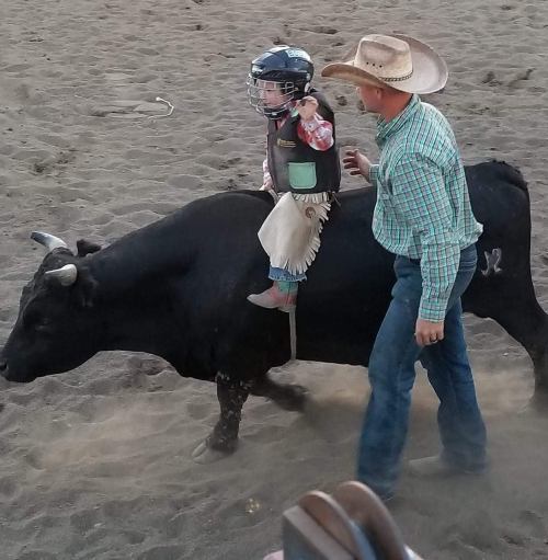 A child in rodeo gear rides a bull while a cowboy guides them in a sandy arena.