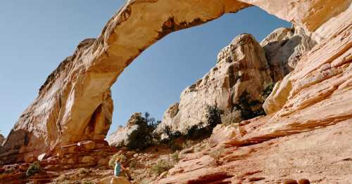 A person sits on a rock beneath a large natural arch in a desert landscape with towering cliffs and clear blue sky.