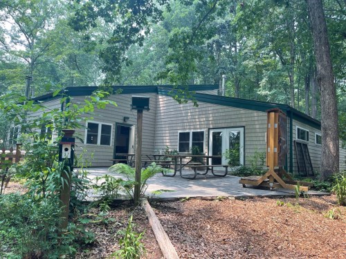 A modern cabin surrounded by trees, featuring a patio with a picnic table and a wooden structure in the foreground.