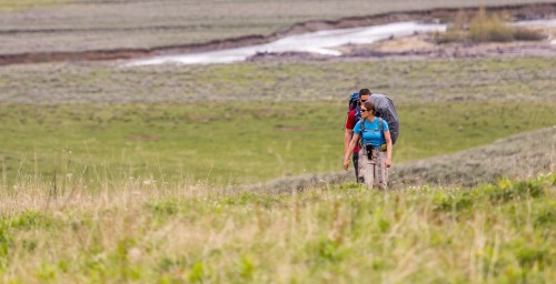 Two hikers ascend a grassy hill, with a stream and lush landscape in the background.