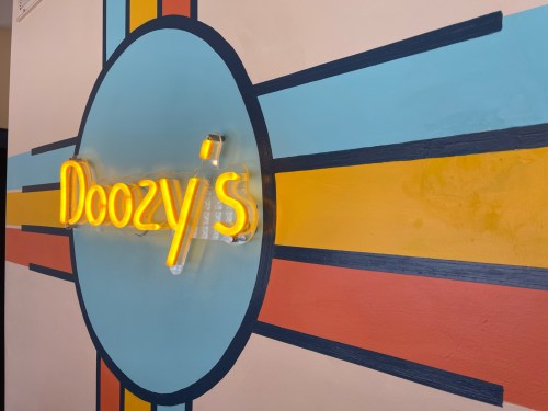 Neon sign reading "Doozy's" on a colorful, retro-style wall with rays of blue, yellow, and red.
