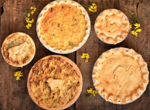 A variety of baked pies on a wooden table, surrounded by small yellow flowers.