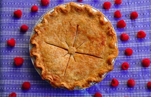A golden-brown pie with a decorative crust, surrounded by fresh raspberries on a blue patterned tablecloth.