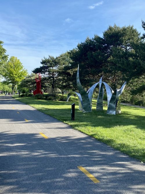 A pathway lined with large metallic sculptures resembling grass, surrounded by trees and blue sky.